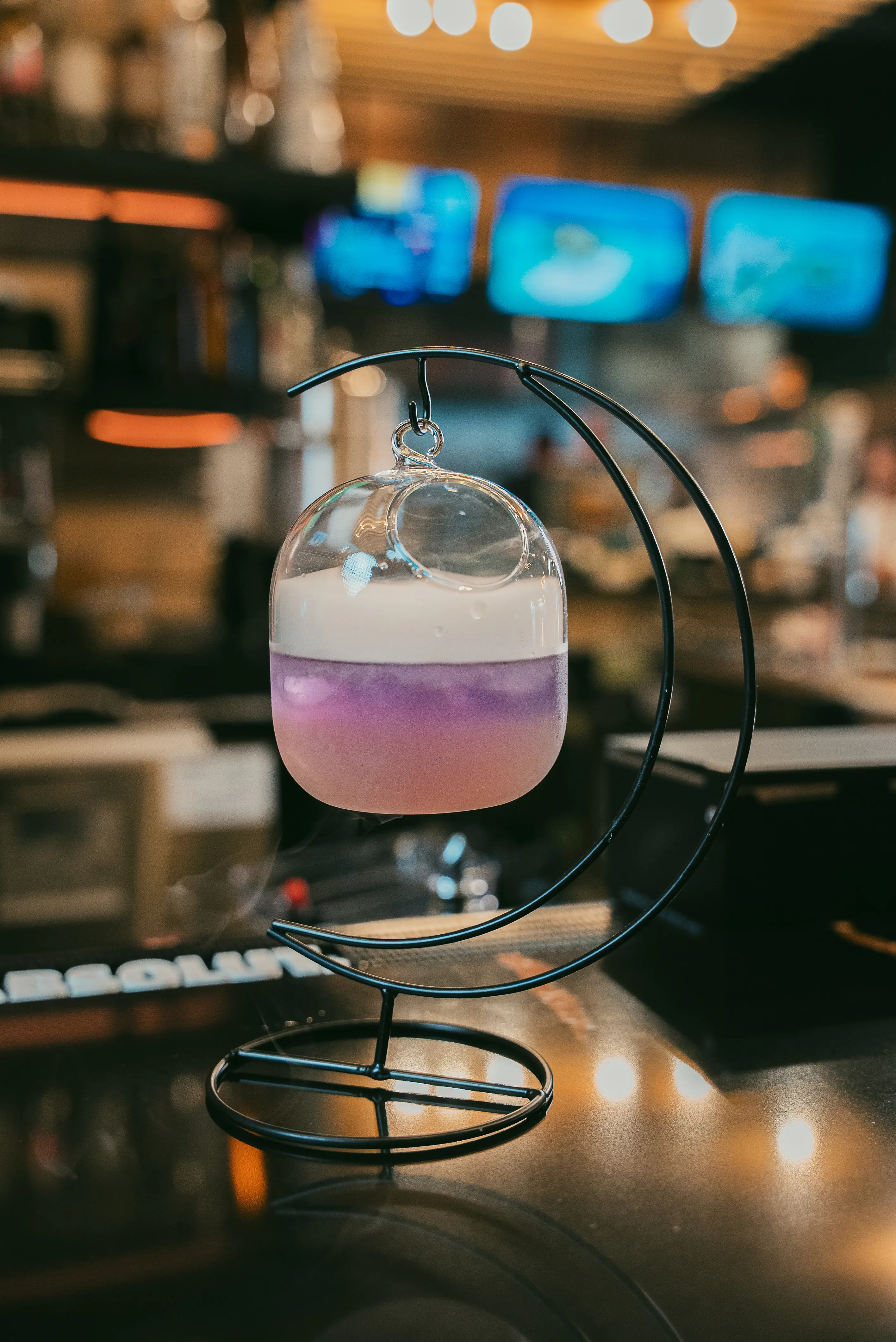 A unique drink presentation featuring a glass sphere with layers of purple and white, elegantly suspended on a black curved stand, set on a bar counter.